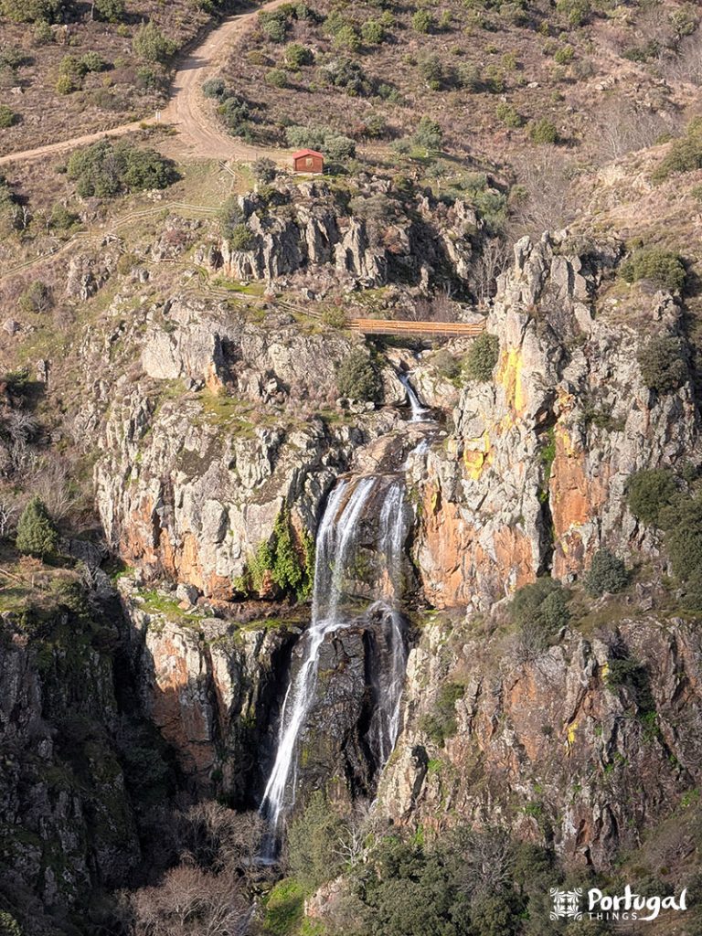 Uma cachoeira alta, Faia d'Água Alta, desce em cascata por penhascos rochosos ao longo da trilha de caminhada Trilho da Cascata. A vegetação esparsa circunda um caminho de terra e uma pequena cabana vermelha no topo. A paisagem parece acidentada e natural.