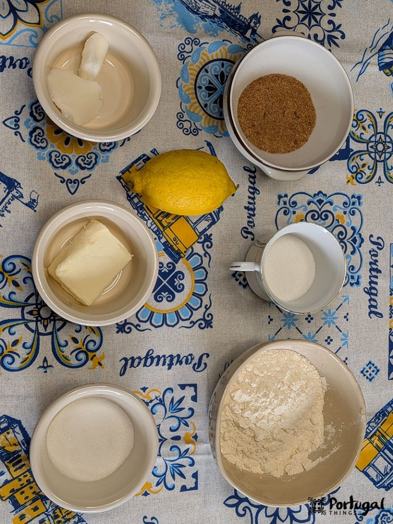 Sete tigelas com ingredientes de panificação (manteiga, açúcar, farinha, açúcar mascavo e banha) e um limão inteiro estão dispostos em uma toalha de mesa decorativa com tema de Portugal, prontos para um Areias de Cascais Receita Fácil. Desenhos em azul e amarelo completam a cena.