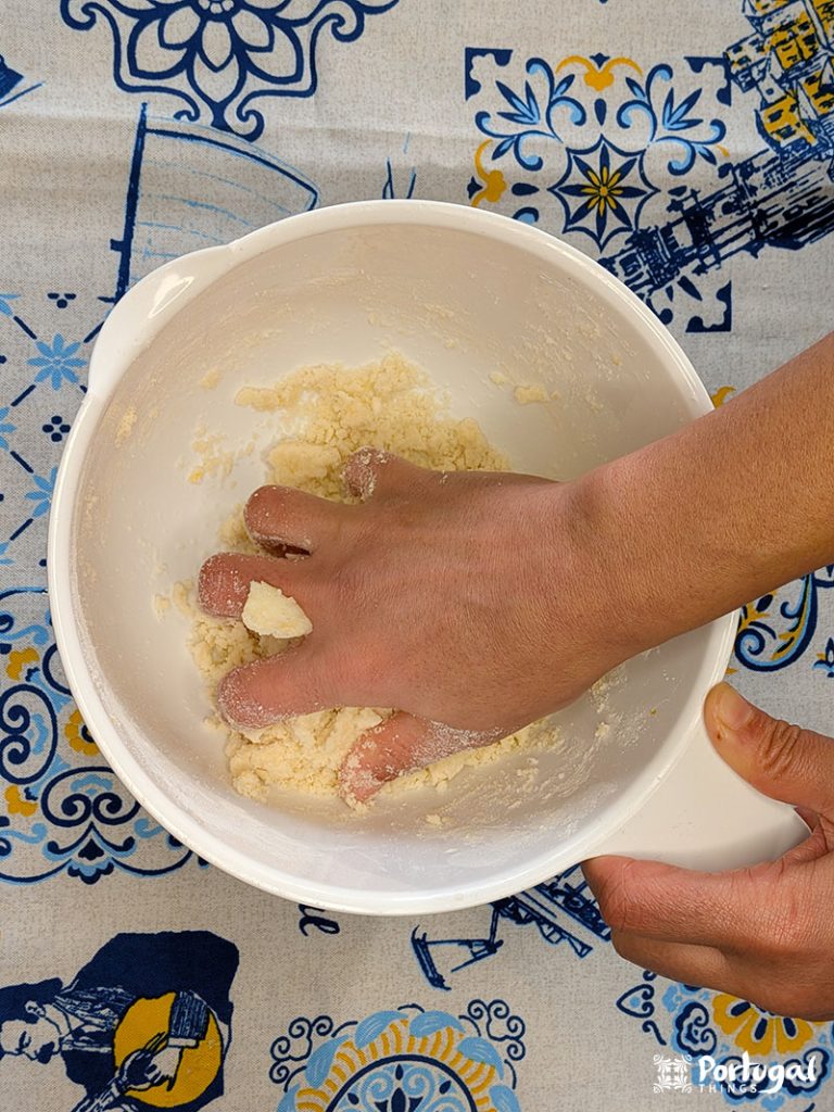 Uma mão mistura a massa em uma tigela branca sobre uma toalha de mesa colorida com desenhos azuis e amarelos, preparando uma receita fácil do Areias de Cascais.