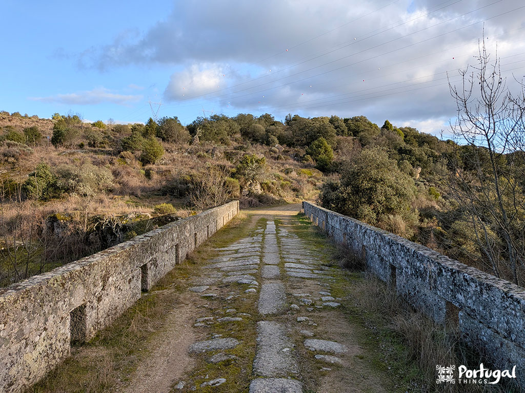 Uma ponte de pedra com paralelepípedos desgastados pelo tempo atravessa uma vala rasa, levando a um trilho pedestre cercado por arbustos e árvores sob um céu parcialmente nublado.
