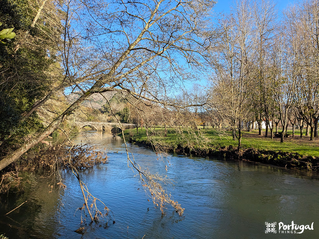 A calm river flows through a park with leafless trees and green grass under a clear blue sky in Caminha. An old stone bridge, part of the Coura Margins Trail.