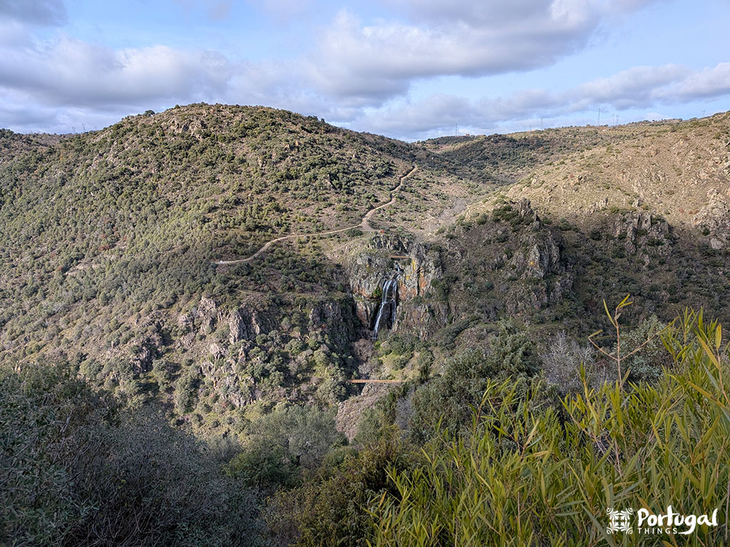 Vista panorâmica de colinas verdes escarpadas com uma estrada sinuosa, uma pequena cachoeira caindo em cascata por penhascos rochosos ao longo da trilha de caminhada do Trilho da Cascata e céu parcialmente nublado.