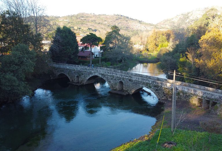 Uma ponte em arco de pedra atravessa um rio calmo cercado por árvores e vegetação ao longo do Trilho nas Margens do Coura, com colinas e casas perto de Caminha visíveis ao fundo sob um céu claro. Cover