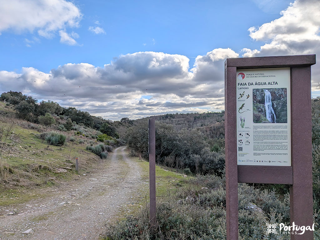 Uma trilha de caminhada de terra serpenteia por uma paisagem montanhosa e verde sob um céu parcialmente nublado. Em primeiro plano, uma placa com texto e imagens fornece informações sobre a cachoeira Faia d'Água Alta e a rota cênica Trilho da Cascata, em Portugal.