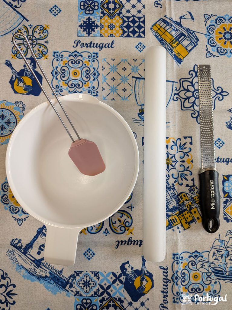 Uma tigela branca com uma espátula rosa fica ao lado de um rolo de massa branco e um ralador com cabo preto, todos dispostos em uma toalha de mesa azul, amarela e branca com tema de Portugal - utensílios perfeitos para preparar o Receita Fácil ou o Areias de Cascais.