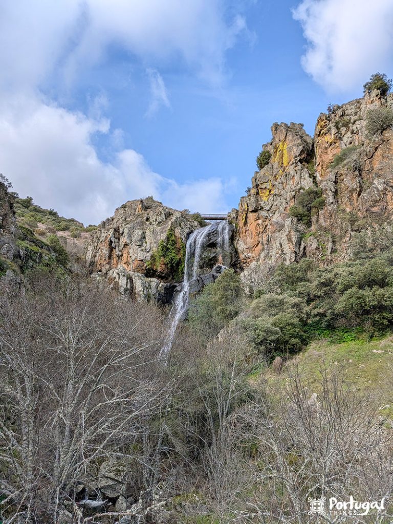 Uma cachoeira alta, Faia d'Água Alta, desce em cascata por penhascos rochosos cercados por árvores esparsas e vegetação sob um céu parcialmente nublado.