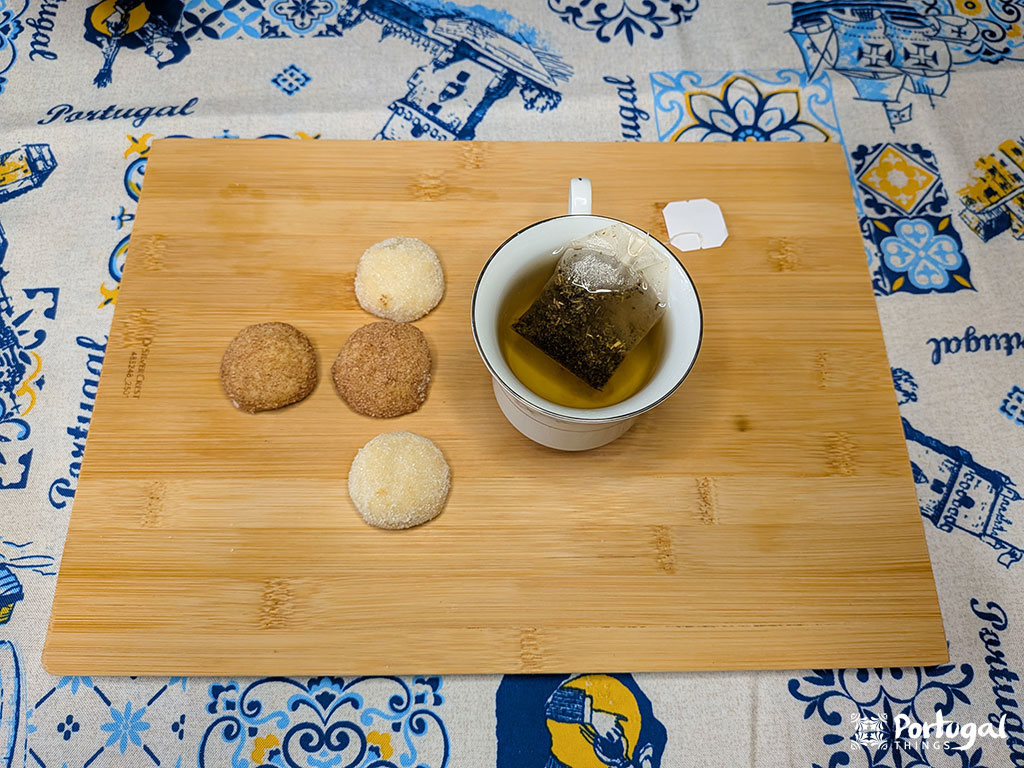 Uma xícara branca com um saquinho de chá fica sobre uma tábua de madeira ao lado de quatro biscoitos redondos Areias de Cascais. A tábua está apoiada em uma toalha de mesa com desenhos azuis e amarelos com a temática de Portugal.