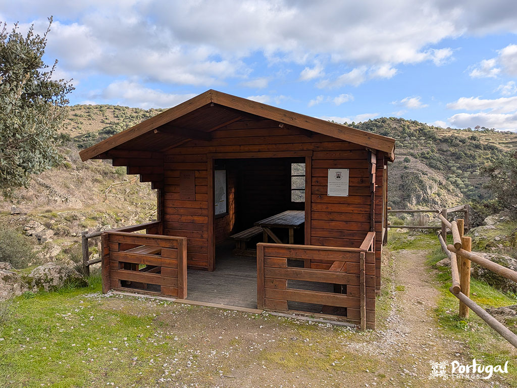 Uma pequena cabana de madeira com uma entrada aberta e uma mesa de piquenique dentro fica na trilha de caminhada do Trilho da Cascata em uma paisagem montanhosa. O céu está parcialmente nublado.