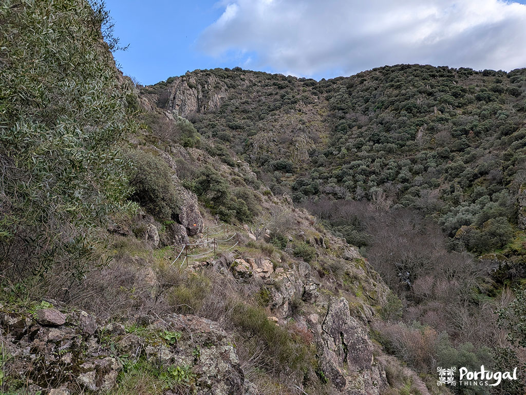 Vista de uma encosta rochosa e acidentada coberta por arbustos verdes ao longo da trilha de caminhada Faia d'Água Alta sob um céu parcialmente nublado; algumas árvores nuas.