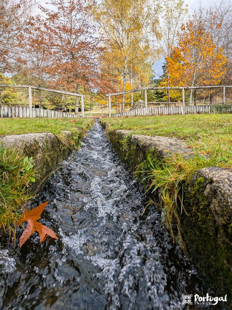 A small stone building with a moss-covered roof sits beside a waterfall in a green, wooded area. The water from the Levada de Víbora flows along a narrow channel that runs past the building, surrounded by lush vegetation and trees.
