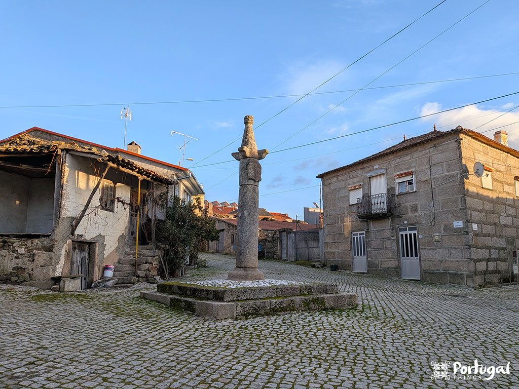 Um pelourinho de pedra fica no centro de uma praça de paralelepípedos, cercado por casas rústicas de pedra sob um céu azul claro - a poucos passos da famosa trilha de caminhada Trilho da Cascata e da deslumbrante Faia d'Água Alta.
