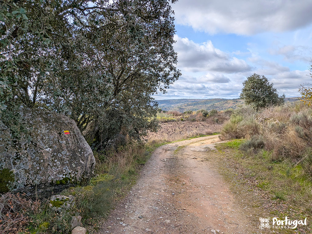 Um caminho de terra serpenteia pela paisagem rural da Faia d'Água Alta, com árvores e arbustos sob um céu parcialmente nublado. Uma grande rocha ao lado da trilha de caminhada exibe um marcador vermelho e amarelo.