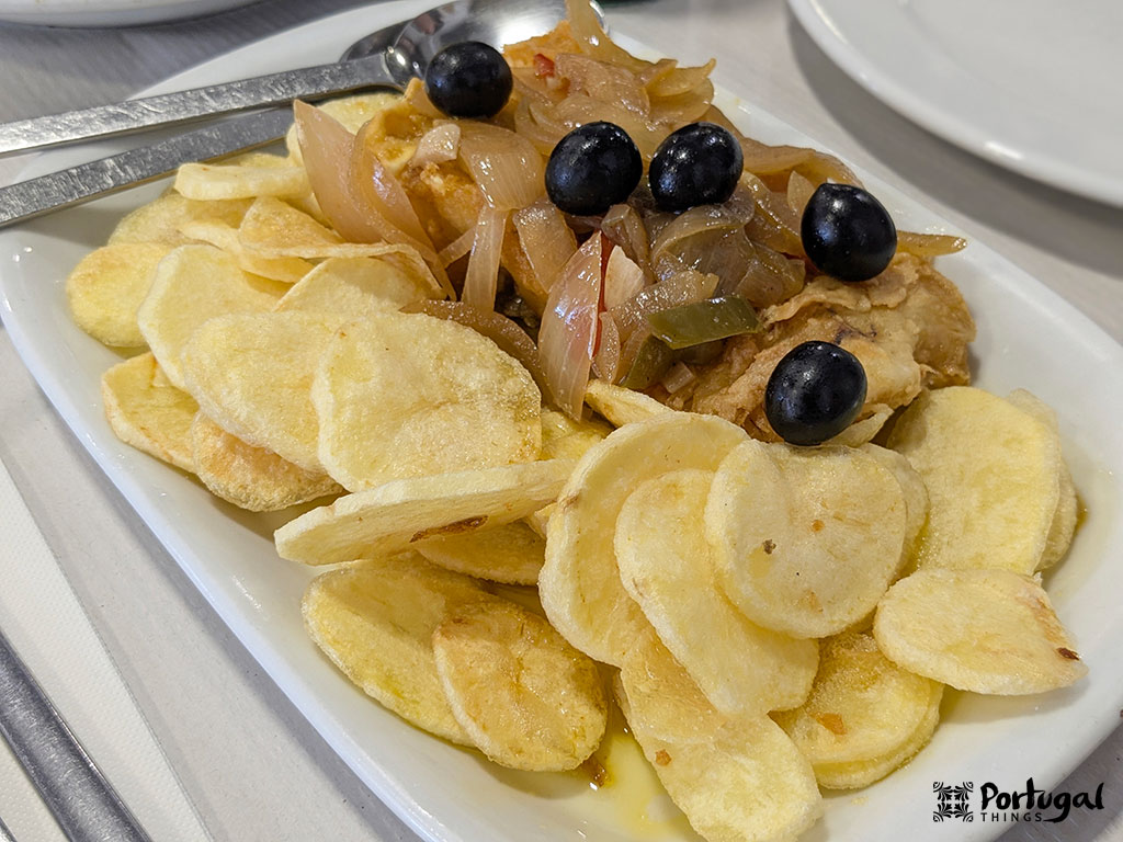 Um prato branco com batatas fritas em fatias finas, cobertas com cebolas cozidas, pimentões, azeitonas pretas e um pedaço de peixe embaixo - um exemplo de comida típica minhota - com utensílios de metal ao lado.