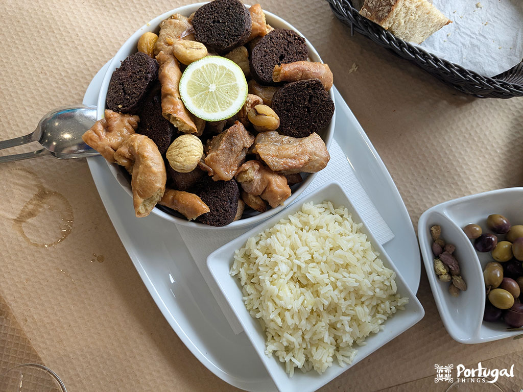 Um prato com arroz branco, uma tigela de carnes variadas, chouriço, castanhas de caju e uma rodela de limão, além de pão, azeitonas e nozes em uma toalha de mesa bege - a verdadeira comida tradicional minhota que destaca os sabores ricos dos pratos do Minho.
