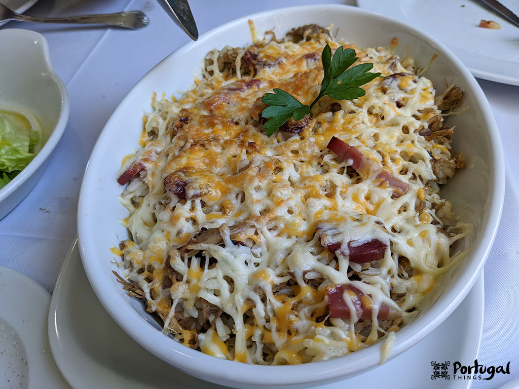 Um prato oval branco cheio de pato desfiado, coberto com queijo derretido e fatias de linguiça - um exemplo convidativo de pratos do Minho - enfeitado com um ramo de salsa, em uma toalha de mesa branca perto de um prato de salada.