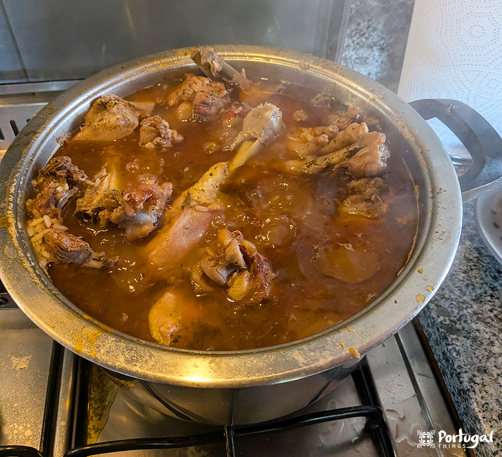 Uma grande panela com pedaços de frango fervendo em um rico ensopado marrom-avermelhado - um exemplo convidativo da culinária portuguesa - fica em cima de uma bancada de granito, com o vapor subindo e o molho borbulhando na borda dessa cena de cozinha aconchegante.