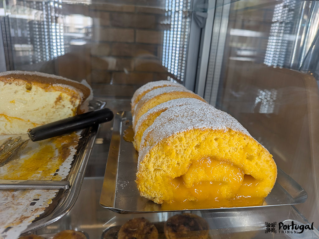 Um close-up de uma tarta de viana recheado com creme de ovo amarelo, polvilhado com açúcar de pasteleiro, exibido em uma bandeja de metal dentro de uma caixa de vidro de padaria - um clássico entre os pratos do Minho.