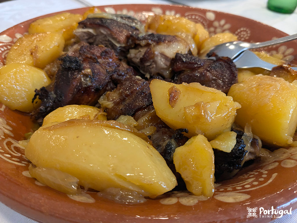 Um prato tradicional português de carne assada e batatas, representando os pratos do Minho, é servido em um prato de barro e guarnecido com cebolas. Um garfo fica ao lado.