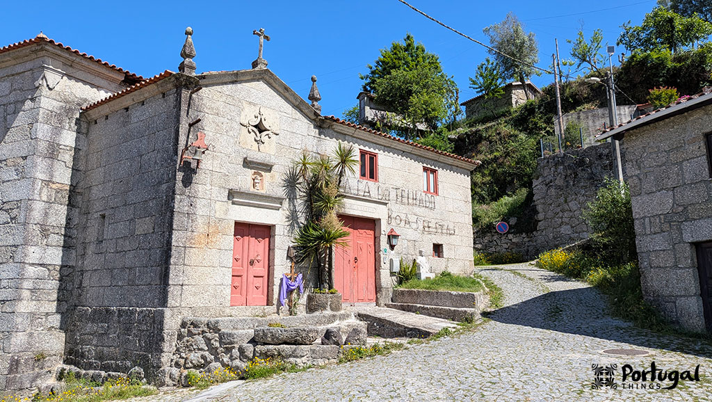 A stone church with red doors and crosses stands at a cobblestone crossroads in a sunny, hilly Portuguese village along the Ave Mills Trail hiking route. Green trees and plants surround the church, with other stone structures nearby.