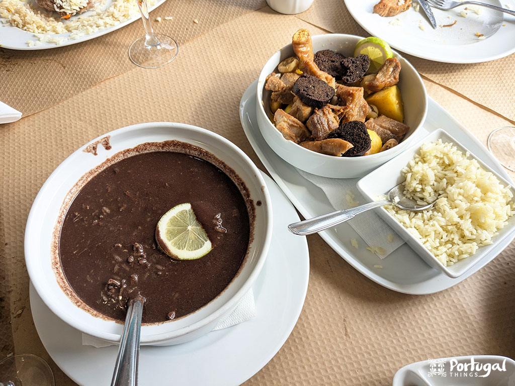 A table set with a bowl of dark soup decorated with a slice of lemon, plates of rice and a fork, as well as mixed meats and sausages - all inviting you to sample the classic flavors of Portugal under a brown paper tablecloth.
