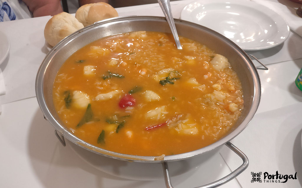 A metal pot filled with Portuguese seafood rice stew, one of Portugal's classic rice dishes, sits on a table alongside bread and white plates – perfect for those wishing to experience the true local flavors.