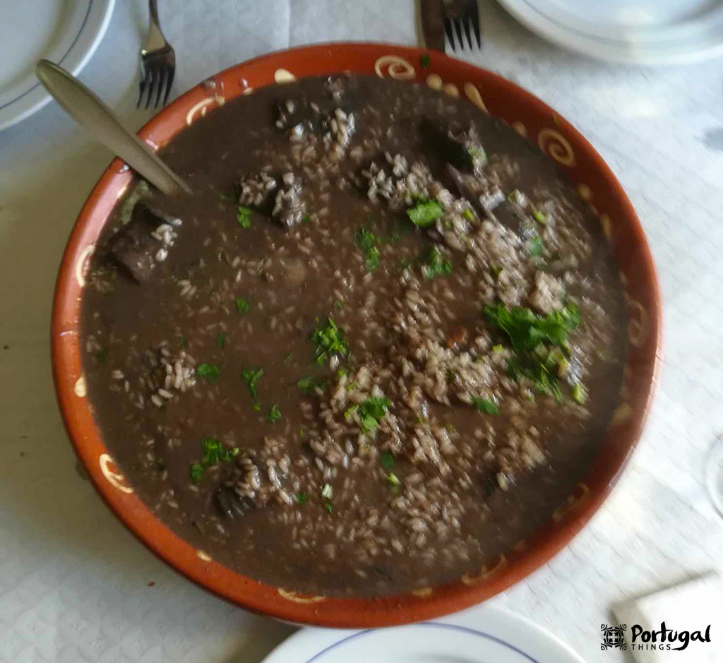 An earthenware bowl filled with sarrabulho rice, one of Portugal's most beloved rice dishes, with dark broth, pieces of meat and fresh herbs, placed on a white tablecloth with plates, cutlery and a spoon ready to be tasted.