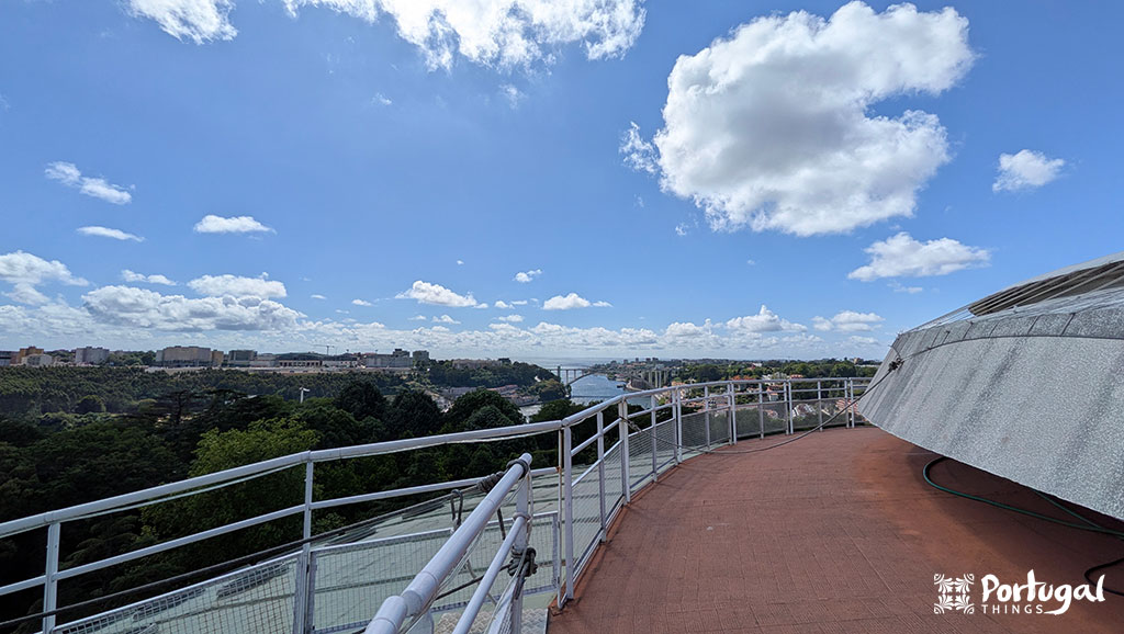 Vista de um terraço na cobertura com grades de metal, com vista para as árvores, o rio e uma ponte distante sob um céu azul com nuvens dispersas. Experimente as vibrações do Porto 360 - vale a pena! 