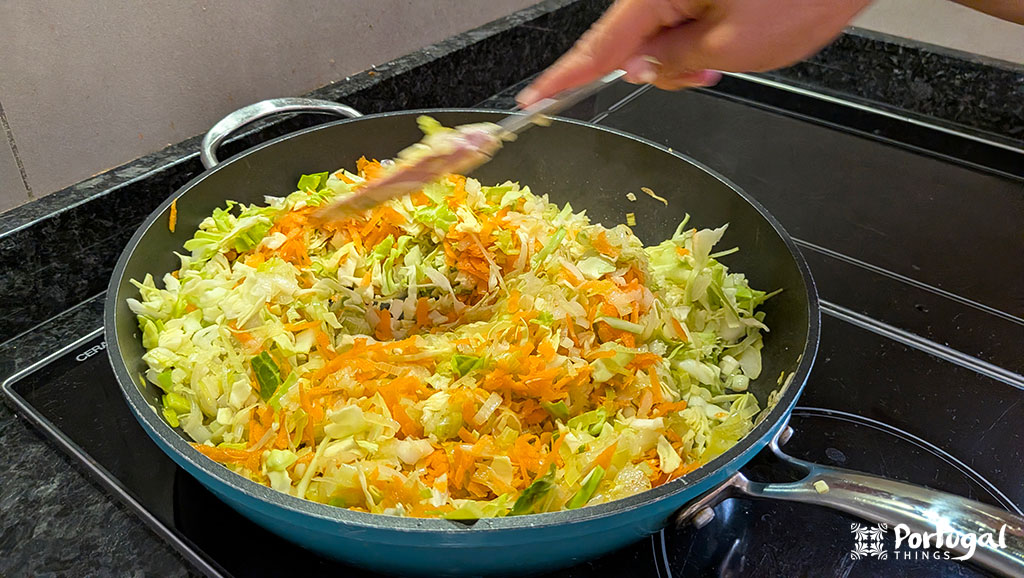 A person stirs shredded cabbage, carrots, and other vegetables in a frying pan on the stovetop, preparing an easy recipe inspired by Legumes à Brás.