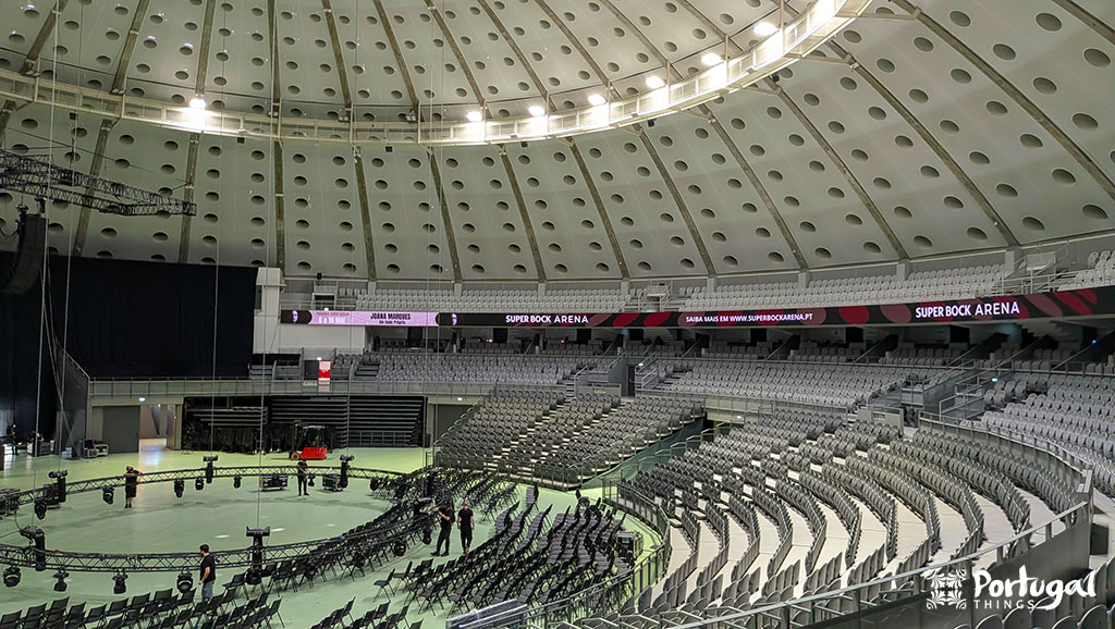 A imagem mostra a Super Bock Arena vazia - antigo Palácio de Cristal - no Porto, com fileiras de assentos cinza em torno de um palco central sob um teto abobadado. Conheça o Porto 360 aqui; vale a pena visitar esse local icônico.