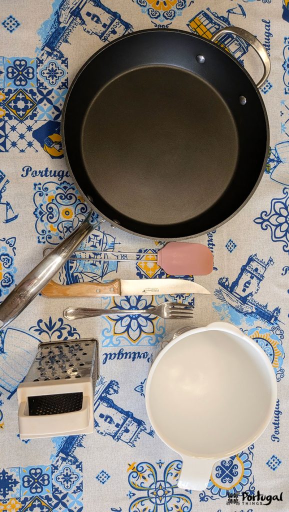 A frying pan, spatula, chef's knife, box grater, and a white mixing bowl arranged on a decorative tablecloth with Portuguese motifs in blue and yellow - perfect for preparing Legumes à Brás or any easy recipe.