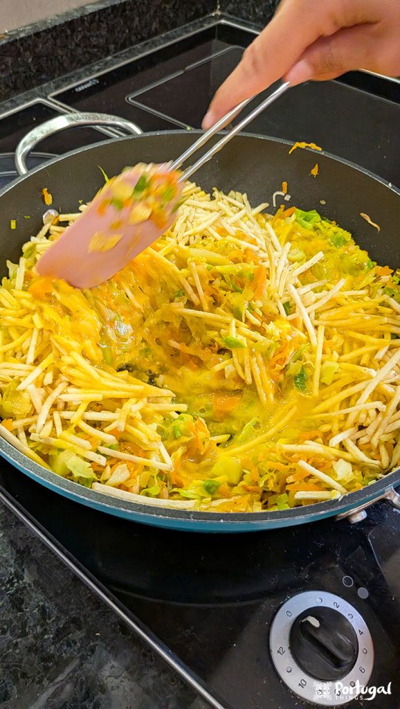 A close-up of a person cooking Vegetables à Brás in a frying pan, mixing shredded vegetables and potatoes on a stovetop with a pink spatula. This easy recipe shows the ingredients being perfectly sautéed.