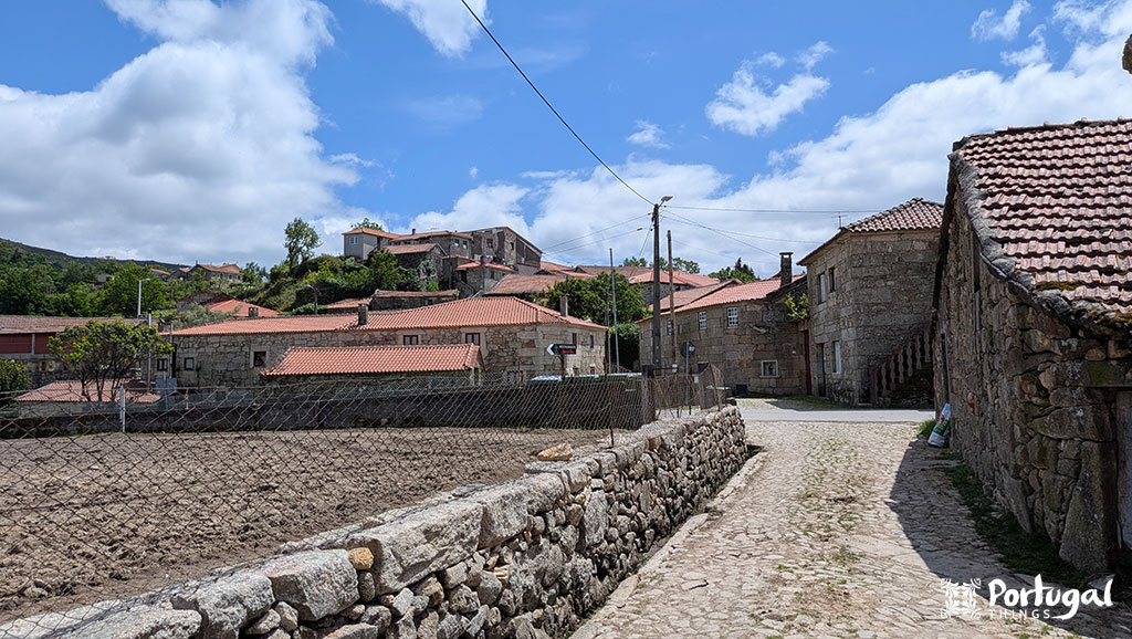 A cobblestone path winds between stone houses with red tiled roofs in a rural Portuguese village on the Ave Mills Trail, a scenic hiking trail for which Portugal is renowned. A plowed field and green hills are visible under a blue sky with scattered clouds.