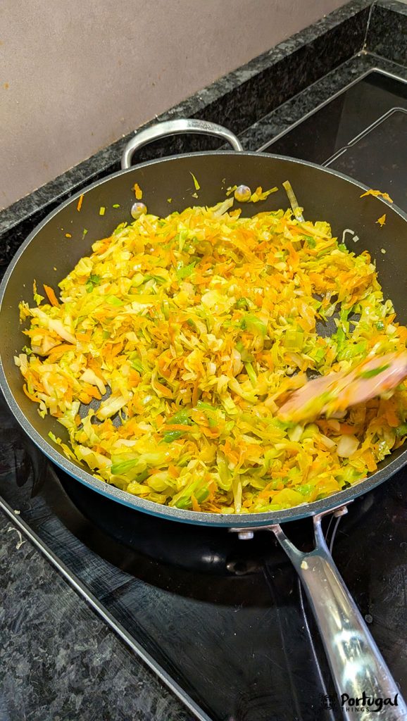 A frying pan on the stove contains grated carrots, cabbage, and sautéed leeks—a true Easy Vegetable à Brás recipe—being stirred with a wooden spatula. The vegetables are lightly cooked and colored.