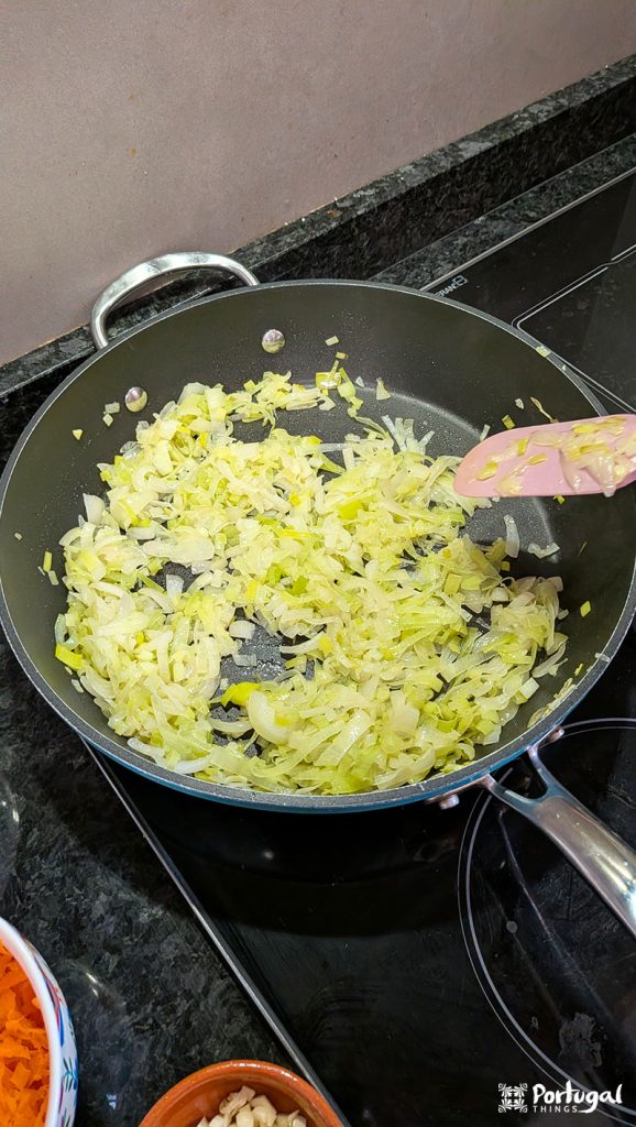 Chopped onions and leeks sautéed in a black pan on the stovetop, stirred with a pink spatula – perfect for preparing Vegetables à Brás, an easy recipe. Other bowls with ingredients can be seen nearby on the countertop.