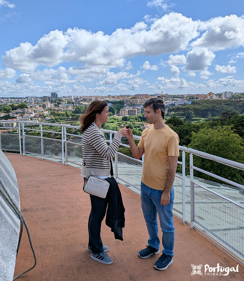 Um homem e uma mulher estão em um mirante panorâmico próximo ao Palácio de Cristal com vista para o Porto 360, de mãos dadas enquanto se encaram sob um céu parcialmente nublado. 
