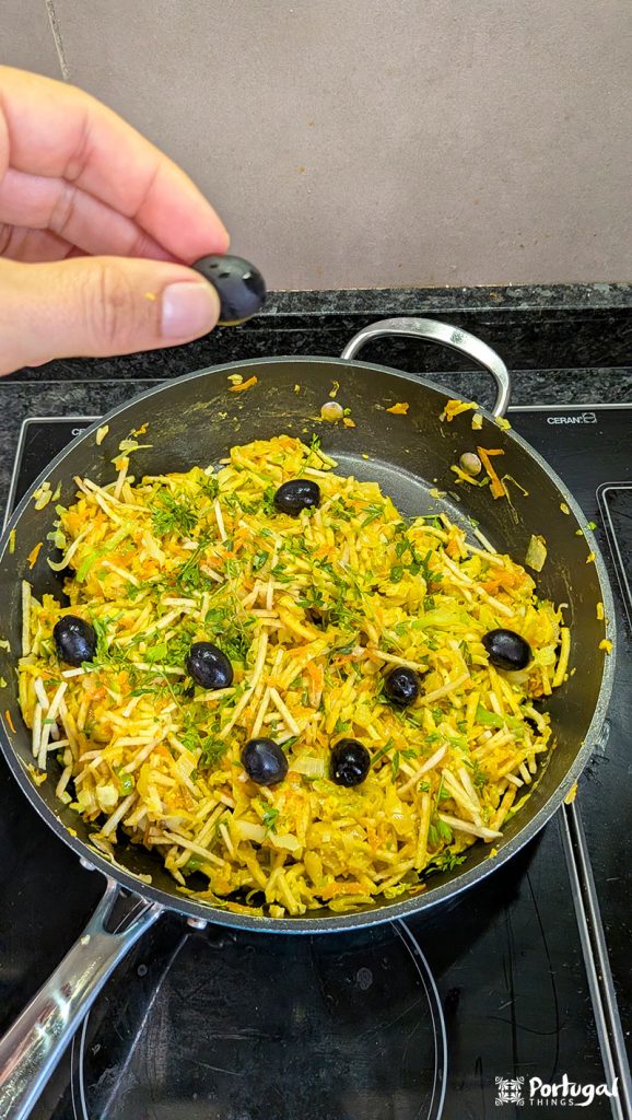A hand holds a black olive over a frying pan full of Legumes à Brás, an easy recipe made with yellow vegetables, herbs, fried potatoes, and more black olives. The dish sits on a stove with a black countertop.