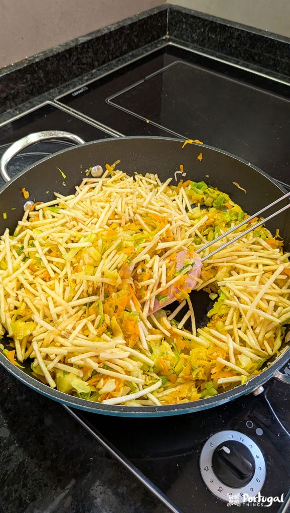 Add shoestring potatoes to the sautéed vegetables in a black frying pan.