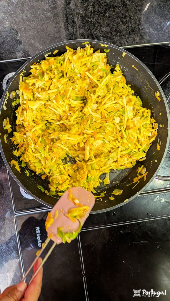 A close-up of a pan full of sautéed and shredded vegetables, probably cabbage and carrots, being stirred with a pink spatula on a black stovetop. This Easy Recipe is inspired by Vegetables à Brás - seasoned and cooked, ready to serve.