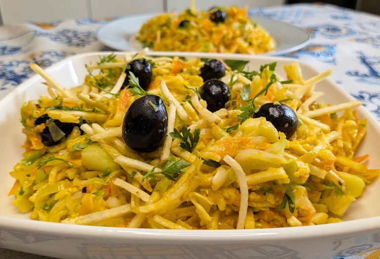 Um close-up de um prato cheio de uma salada colorida com legumes, azeitonas pretas, legumes desfiados e ervas frescas - uma receita fácil e irresistivelmente fresca inspirada no Legumes à Brás, com outro prato desfocado ao fundo.