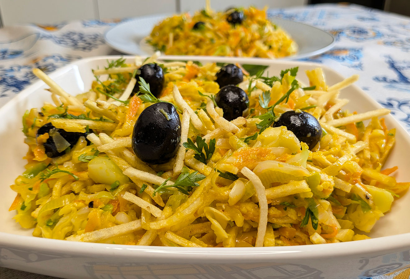 Um close-up de um prato cheio de uma salada colorida com legumes, azeitonas pretas, legumes desfiados e ervas frescas - uma receita fácil e irresistivelmente fresca inspirada no Legumes à Brás, com outro prato desfocado ao fundo.