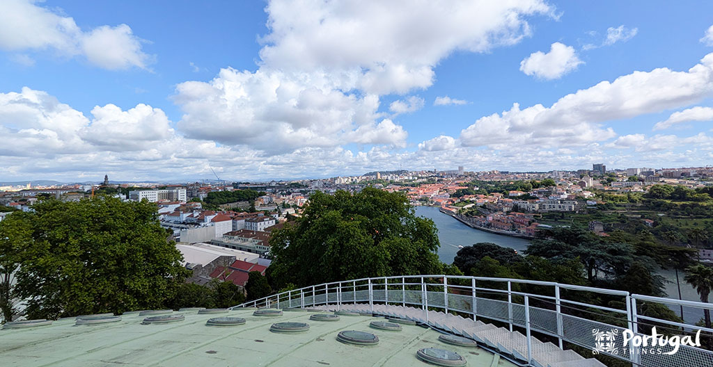 Uma vista panorâmica do rio que serpenteia pelo Porto, com muitos edifícios, árvores verdes e céu parcialmente nublado. A grade curva e o telhado em primeiro plano sugerem uma experiência Porto 360.