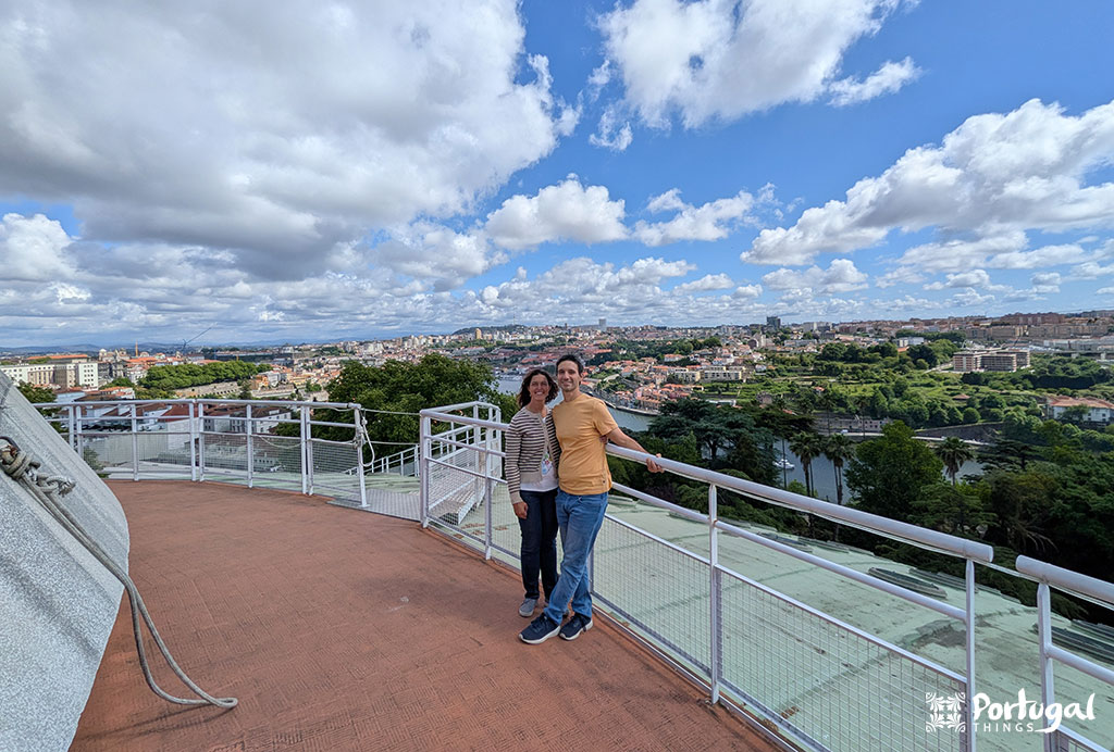 Um casal sorridente está em um deck de observação no telhado com uma vista panorâmica de Lisboa, Portugal, sob um céu parcialmente nublado. Colinas verdes e prédios da cidade se estendem ao fundo - uma experiência que realmente vale a pena. O logotipo 