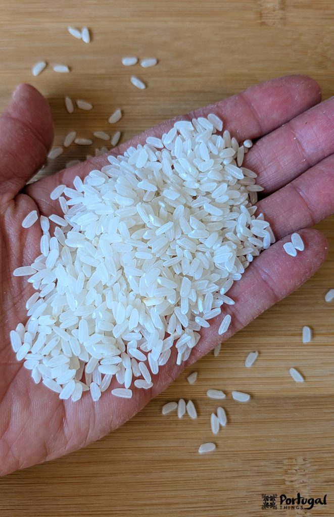 Um close-up de uma mão segurando um punhado de grãos de arroz branco cru, destacando o “arroz português tradição”, com mais arroz espalhado sobre uma superfície de madeira ao fundo.