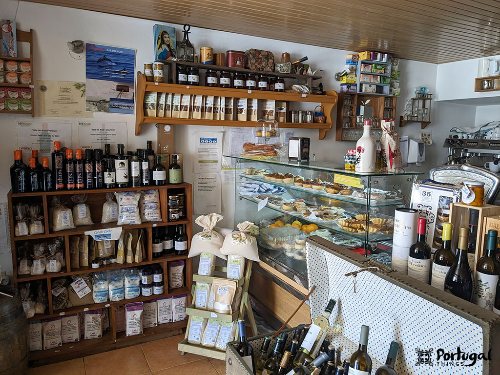 O interior de uma pequena loja com prateleiras repletas de vinhos, produtos embalados e produtos locais. Uma vitrine de vidro exibe doces e sobremesas. Aconchegante e bem organizada, a loja oferece itens gourmet, sabores regionais e variedades de arroz, como o autêntico arroz português.