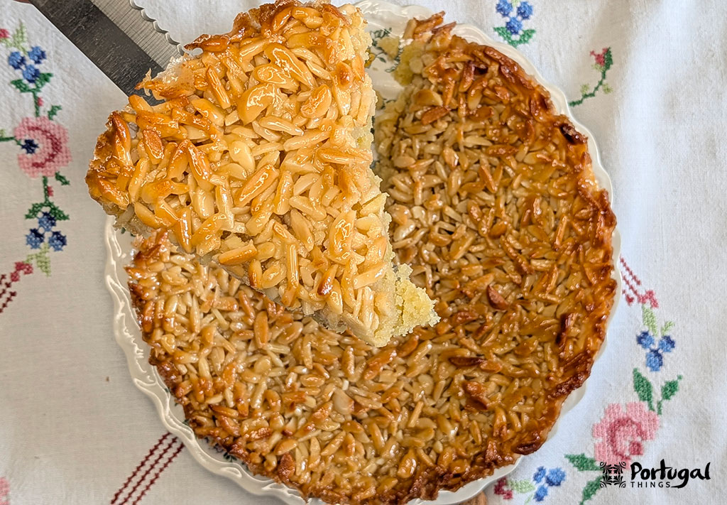 A knife holds a slice of almond tart; beneath it lies a whole almond tart.