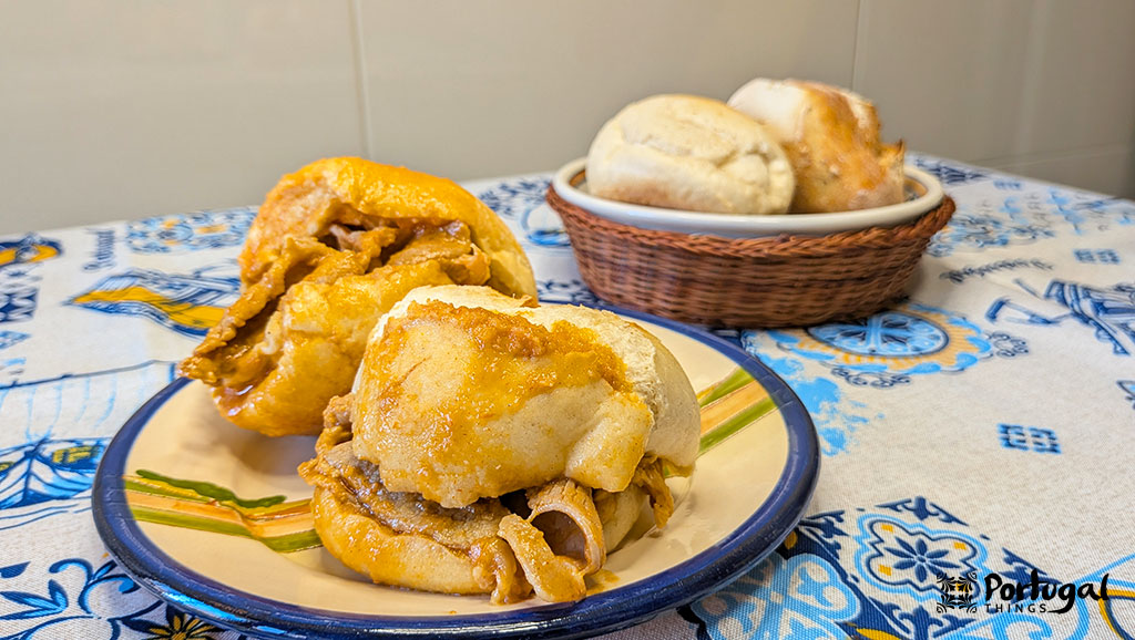 A plate with two juicy bifanas, with another plate in the background holding two bread rolls.