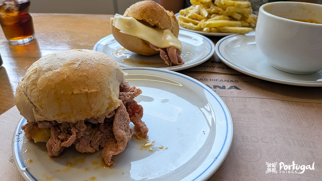 uma mesa 2 bifanas do Porto uma com queijo outra simples um prato de batata frita e um pires com sopa
