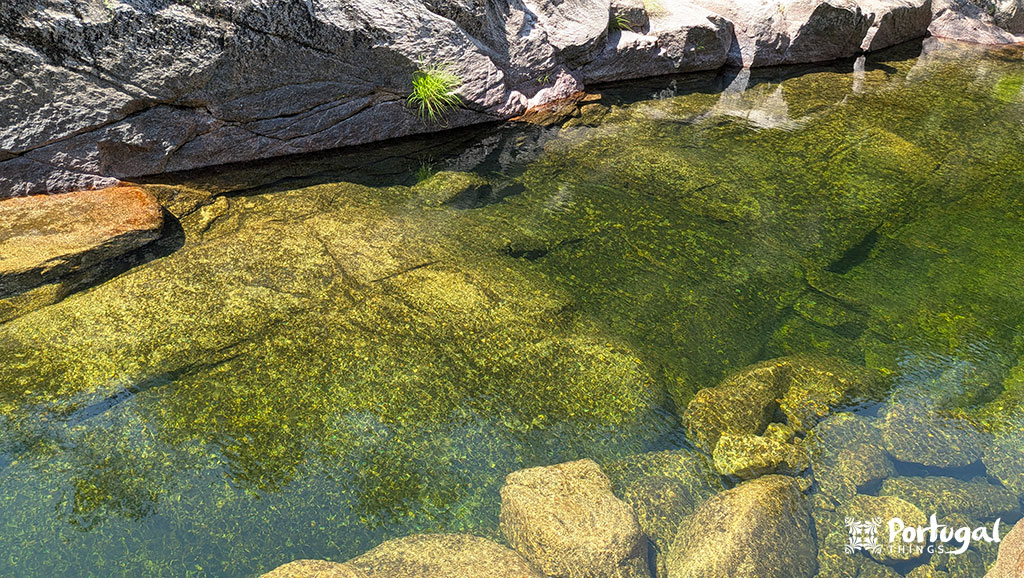 Piscina de águas límpidas, com rochas visíveis sob a superfície, rodeada por rochas maiores. O sol projeta reflexos na água.
