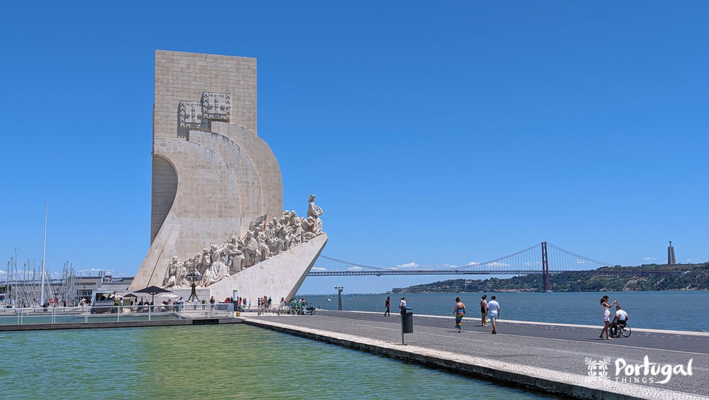 18 best things to do in Lisbon in 3 days - Itinerary 13 um monumentos dedicado aos descobrimentos no rio ao fundo tem uma ponte vermelha e há algumas pessoas a caminhar ao lado no rio